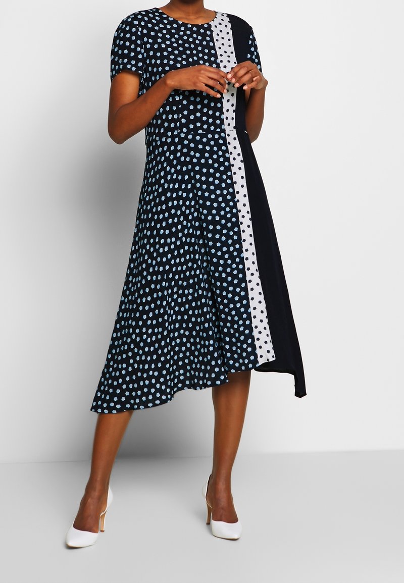 Femme portant une robe midi noire et bleue à motifs avec manches courtes, bande à pois blanche et chaussures blanches à talons hauts, debout sur un sol gris.