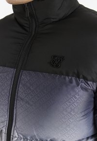 Detailní záběr na černou a fialovou pufrovou bundu SikSilk se zipem a logem SikSilk na hrudi.