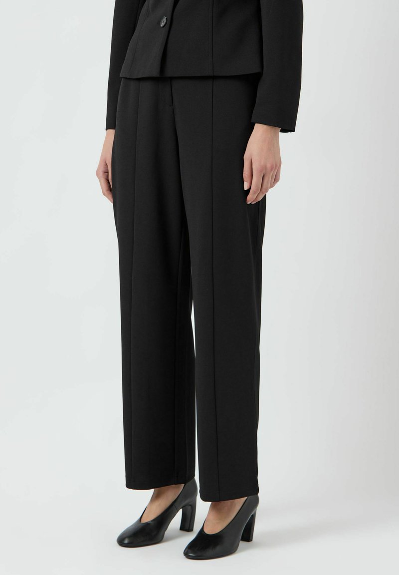 Černé přizpůsobené kalhoty vyrobené ze strukturované tkaniny, charakterizované rovně střiženými nohavicemi, bočními kapsami a hladkou texturou. V kombinaci s černými podpatekmi.