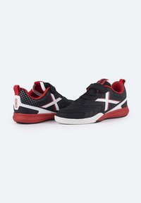 Zapatillas deportivas negras con acentos rojos, que presentan un logo X blanco, una correa de velcro y un patrón de puntos en el talón. Superficie texturada, suela de goma.