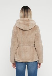Veste à capuche en fausse fourrure beige clair avec une taille cintrée, des manches longues et une surface texturée. Portée sur un jean bleu.
