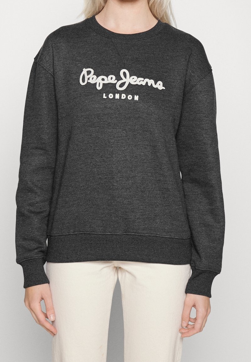 Personne portant un sweat-shirt gris foncé avec le texte blanc « Pepe Jeans London » sur la poitrine, associé à un pantalon clair.