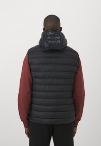 Chaleco acolchado negro con un cuello alto, que presenta secciones acolchadas y una textura suave y brillante, llevado sobre una prenda de manga larga color burdeos.