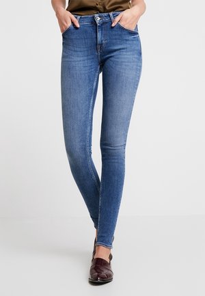 Kobieta ubrana w niebieskie obcisłe dżinsy i brązowe mokasyny, z rękami w kieszeniach, stoi z skrzyżowanymi nogami na jednolitym jasnym tle.