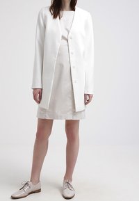 Vit knälång blazer med en knapp, buren över en beige klänning. Outfiten kombineras med ljusa slips- på skor.