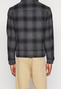 Man med en grå och svart rutiga jacka med knäppte manschetter och beige byxor, visad bakifrån mot en neutral bakgrund.