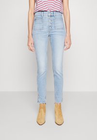 Person wearing high-waisted light blue jeans with front buttons and tan suede ankle boots, standing against a plain light background.