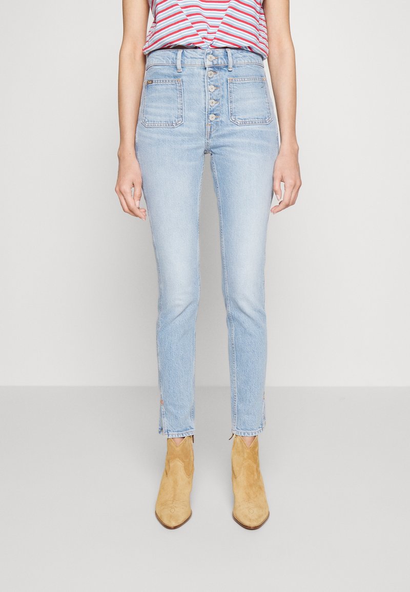 Person wearing high-waisted light blue jeans with front buttons and tan suede ankle boots, standing against a plain light background.