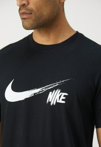 Black cotton t-shirt featuring a large white Nike swoosh logo and distinctive brushstroke text. Crew neck design, short sleeves.