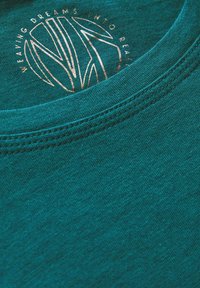 Teal katoenen t-shirt met een kraag met decoratieve stiksels, met een rond logo waarop staat "DROMEN WEVEN TOT REALITEIT." Een gladde textuur.