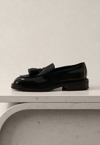Μαύρο δερμάτινο παπούτσι μοκασίνι με φούντες σε ανοιχτό μπεζ μαρμάρινη επιφάνεια με καμπύλη λεπτομέρεια ακμής.