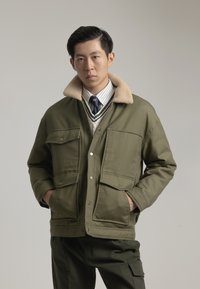 Chaqueta de color verde oliva con un cuello de pelaje sintético beige, dos bolsillos frontales, cierre de botón a presión y un diseño de tela texturizada.