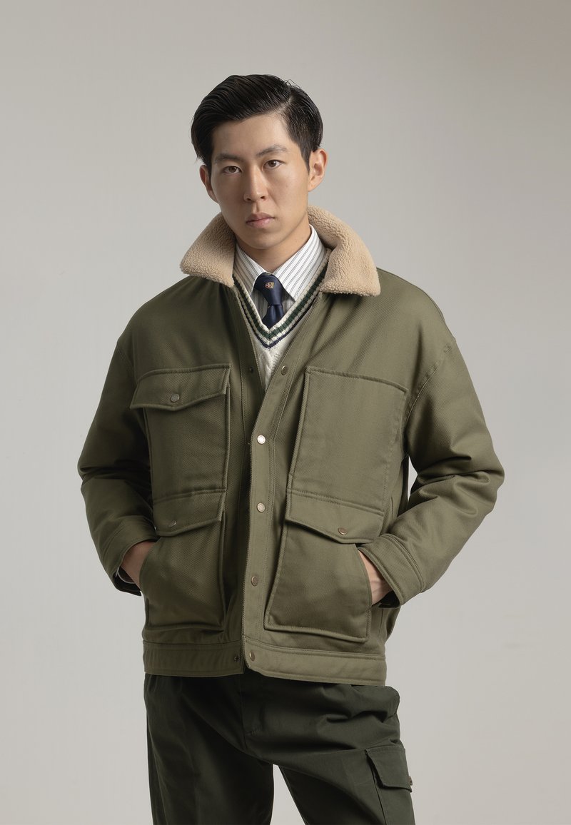 Chaqueta de color verde oliva con un cuello de pelaje sintético beige, dos bolsillos frontales, cierre de botón a presión y un diseño de tela texturizada.
