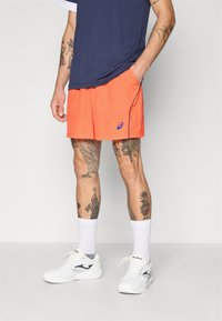 Pomarańczowe spodenki sportowe z bocznymi kieszeniami i niebieskim logo, zestawione z granatową koszulą i białymi sneakersami, z widocznymi tatuażami na nogach.