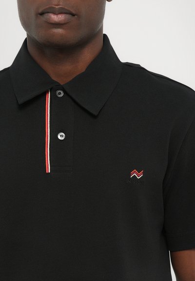 Polo noir en tissu texturé avec col à boutons, présentant une rayure rouge et blanche sur la patte de boutonnage et un petit logo brodé.