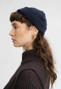 Marineblauwe gebreide muts met een omgeslagen braak, gedragen door een persoon met krullend haar, gecombineerd met een gestructureerde bruine trui met hoge kraag.