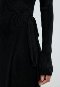 Schwarzes geripptes Kleid mit langen Ärmeln, Wickeldesign und einer Bindeschleife in der Taille. Weicher Stoff mit figurbetonter Silhouette.