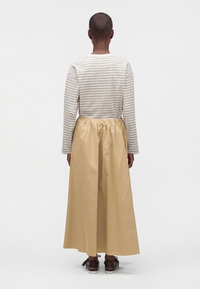 Personne debout de dos portant une longue jupe beige, un haut rayé à manches longues et des chaussures marron foncé à lacets sur fond blanc.