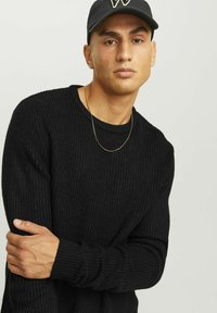 Maglione nero lavorato a maglia con una texture a coste, scollatura rotonda e maniche lunghe. Collana in catena dorata e berretto nero con logo.