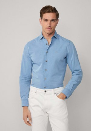 Hombre vestido con una camisa abotonada azul claro y pantalones blancos, de pie con una mano en el bolsillo contra un fondo liso.