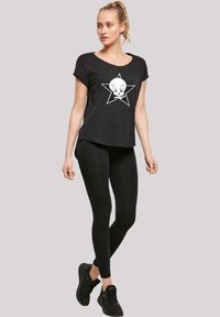 Svart t-skjorte med oppvikte ermer og hvit Tweety Bird-grafikk inni en stjerne, kombinert med svarte leggings og svarte joggesko.