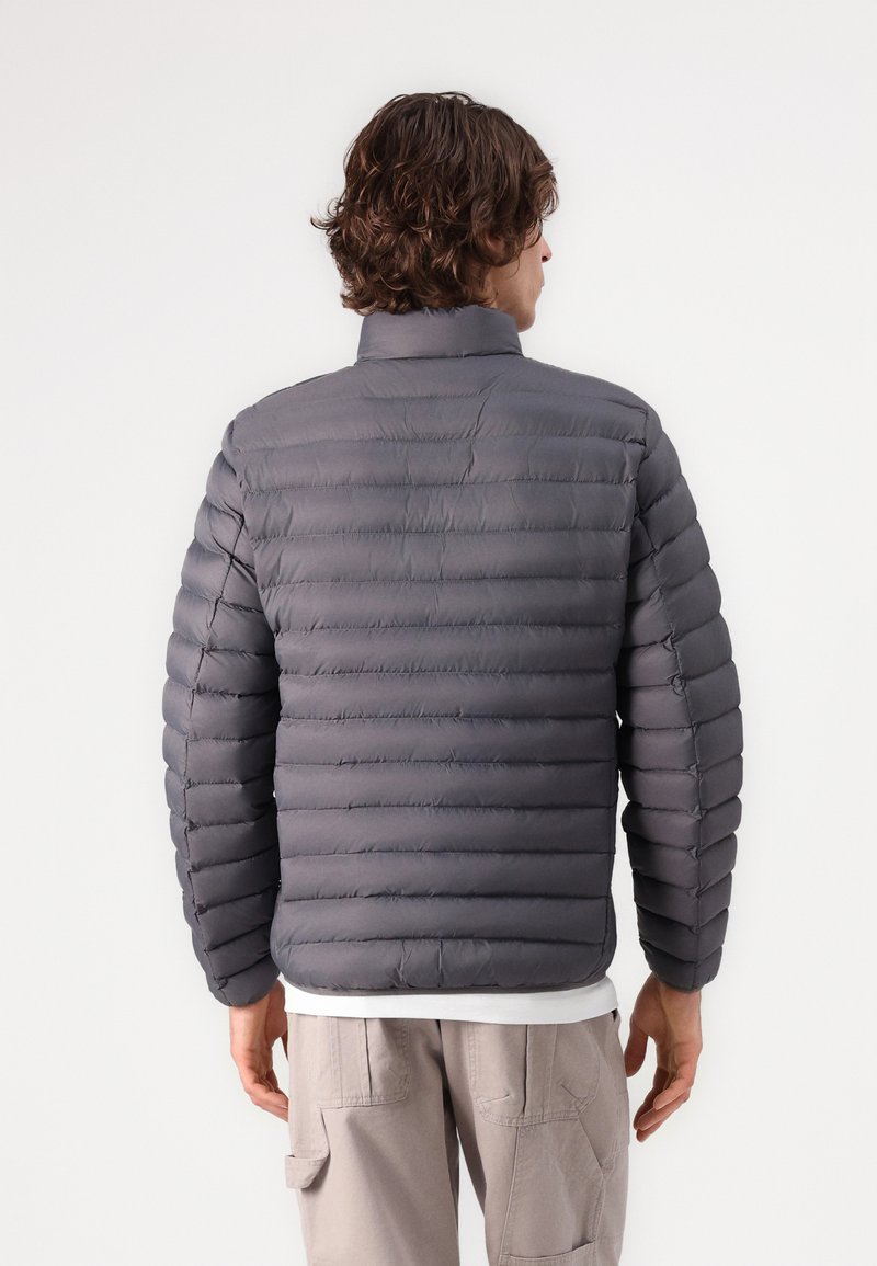Homme aux cheveux bruns ondulés portant une veste grise matelassée et un pantalon cargo beige, tourné le dos sur un fond blanc uni.
