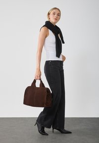 Bruine suede handtas met dubbele handvaten, een gestructureerde vorm en tonale stiksels. Model draagt een witte tanktop en een zwarte sjaal, gecombineerd met zwarte jeans.
