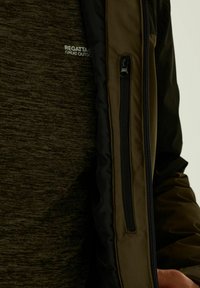 Primer plano de una persona que lleva una camiseta Regatta Great Outdoors de color verde oscuro y una chaqueta verde oliva con un bolsillo vertical con cremallera.