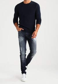 Marinblå texturerad stickad tröja kombinerad med slitna blå jeans och vita sneakers; jeansen har revor och urblekta detaljer för en avslappnad look.