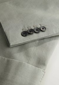 Manche en tissu gris clair texturé avec quatre boutons gris foncé cousus en ligne près du poignet, montrant des coutures détaillées.