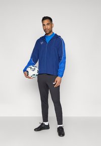 Μπλε και ναυτικό αθλητικό μπουφάν με φερμουάρ και κουκούλα, συνδυασμένο με μπλε πουκάμισο, γκρίζα παντελόνια και μαύρα ποδοσφαιρικά παπούτσια; κρατώντας μια μοτίβο ποδοσφαιρική μπάλα.