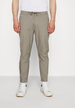 Mies, jolla on päällään kapealahkeiset beiget housut, valkoiset sukat ja valkoiset lenkkarit, seisomassa yksinkertaisen valkoisen taustan edessä.