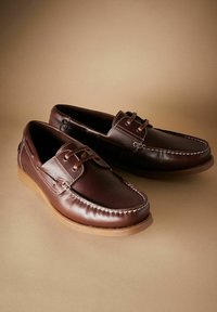 Braune Lederbootsschuhe mit einer glatten Oberfläche, die weiße Nähte, Schnürakzente und eine Gummisohle aufweisen. Sie haben ein rundes Zehendesign.
