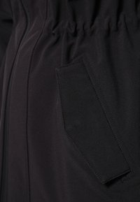 Vêtement d'extérieur noir avec un tissu texturé, une taille élastiquée froncée et une poche latérale dissimulée.