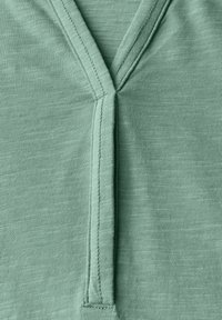 Top en coton à col en V de couleur vert clair, avec une finition texturée et un subtil détail de couture le long de l'encolure et de la patte de boutonnage.