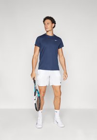 Camiseta desportiva azul-marinho, calções brancos e sapatilhas brancas. A segurar uma raquete de ténis com um destaque azul. Branding mínimo visível.