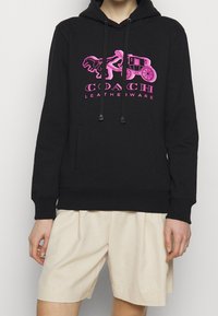 Sudadera negra con capucha que presenta un logo gráfico en rosa de un caballo y carruaje con el texto "COACH LEATHERWARE", hecha de un tejido suave y con un bolsillo frontal.