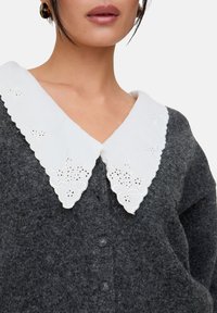 Graue Wollstrickjacke mit großem weißem, besticktem Kragen, der gewellte Ränder und Blumenmuster aufweist; Knopfleiste vorne.