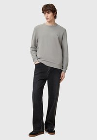 Sudadera gris claro con cuello redondo, puños acanalados y logo en el pecho, combinada con jeans de pierna ancha oscuros y zapatos negros. Fondo liso.