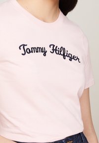 Camiseta de algodón rosa con texto bordado en azul marino "Tommy Hilfiger". Mangas cortas y cuello redondo clásico. Textura suave.