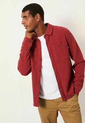 Red textured shirt with a buttoned front, two chest pockets, and a collared neckline, worn over a white T-shirt and khaki pants.