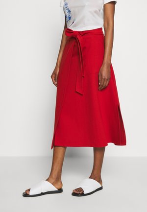 Femme portant une jupe midi portefeuille rouge avec des fentes latérales et une ceinture nouée à la taille, associée à des sandales blanches à enfiler et un t-shirt blanc à motif floral.