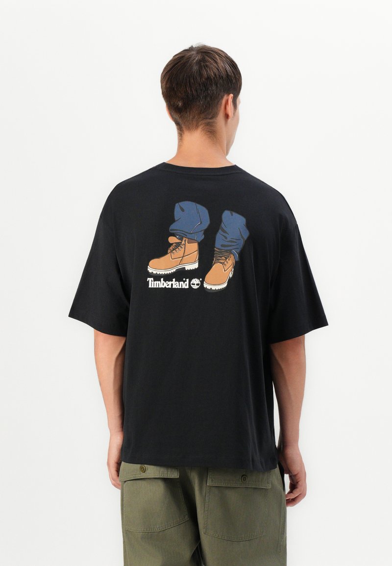 Maglietta nera con grafica di stivali e jeans sul retro. Vestibilità ampia, colletto rotondo e realizzata in tessuto di cotone.