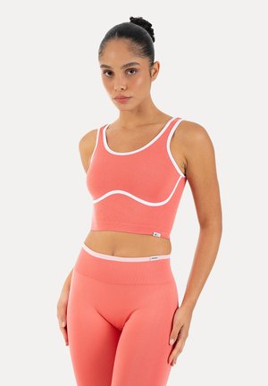 Kvinde iført koral pink tætsiddende crop top og højtaljede leggings med hvid kant, stående mod en ensfarvet lys baggrund.