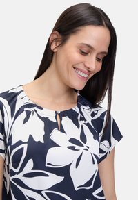 Navy bluse med store hvide blomsterprint, rund halsudskæring med knap-detail og korte ærmer. Glat stofstruktur.