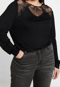Schwarzes Langarmoberteil aus transparenter Mesh mit gewelltem Design am Ausschnitt, kombiniert mit dunkelgrauen Jeans. Glatte Stoffoberfläche.