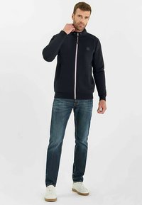 Marineblauw jack met rits en hoge kraag, roze rits en zijzakken, gecombineerd met een blauwe spijkerbroek en witte sneakers. Gladde stofstructuur.