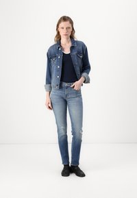 Denim jacket in dark blue with two front pockets and button closure, paired with fitted, light blue distressed jeans and black shoes.
