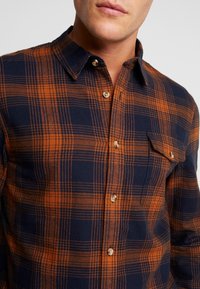 Camisa de franela en cuadros azul marino y naranja, con un diseño de botones, cuello puntiagudo y un bolsillo en el pecho. Incluye botones redondos.