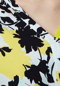 Korte mouwen blouse met een bloemenpatroon in zwart, geel en lichtblauw op een witte achtergrond. Beschikt over een V-hals en een soepele textuur.